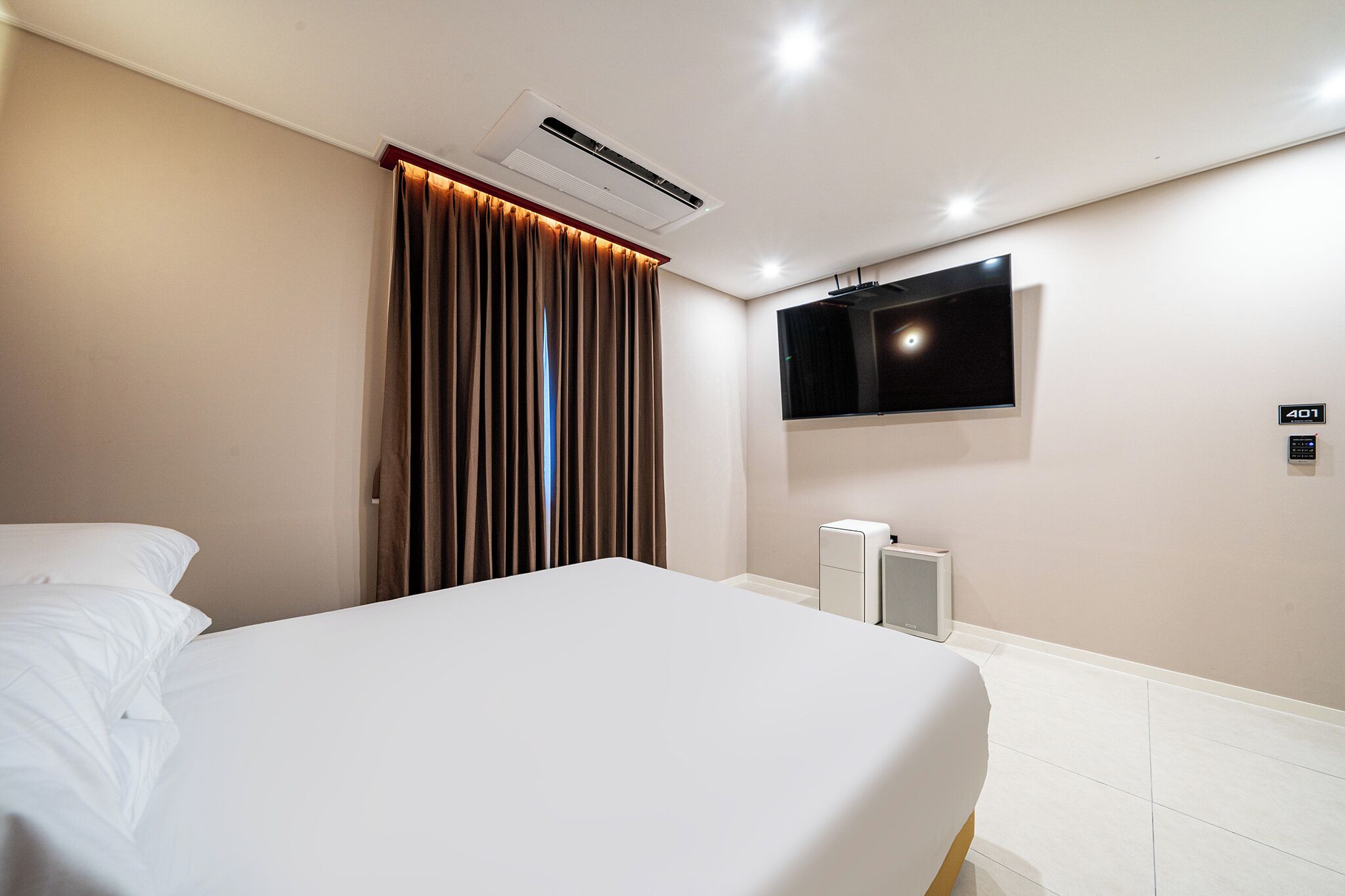 A hotel room with a bed, an air conditioning unit, a television mounted on the wall, and a control panel.