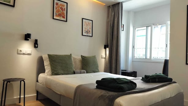 Een moderne hotelkamer met een bed, groene kussens, een klein krukje en kunst aan de muur.