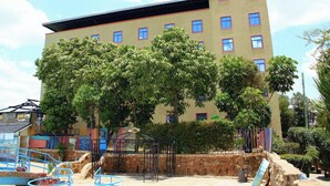 A multi-story building with a flat roof, surrounded by lush green trees and a playground area with blue equipment. - Global Hotels & Suites Kitengela (Kitengela)