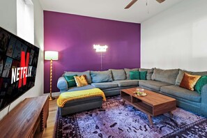 Een woonkamer met een hoekbank, een salontafel, een aan de muur bevestigde televisie en een vloerlamp.