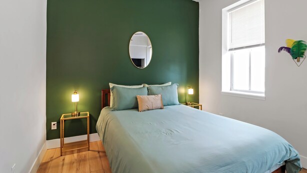 Quarto com cama, duas luminárias de cabeceira, um espelho redondo e uma janela com persianas.