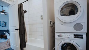 Uma área de lavanderia com máquina de lavar e secadora, um chuveiro com cortina, e uma porta que dá para uma sala de estar com sofá e ventilador de teto.