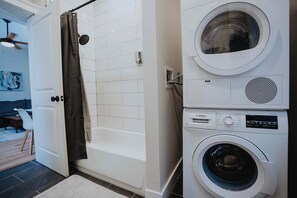 Un área de lavandería con lavadora y secadora, una ducha con cortina, y una puerta que da a un espacio de estar con sofá y ventilador de techo.