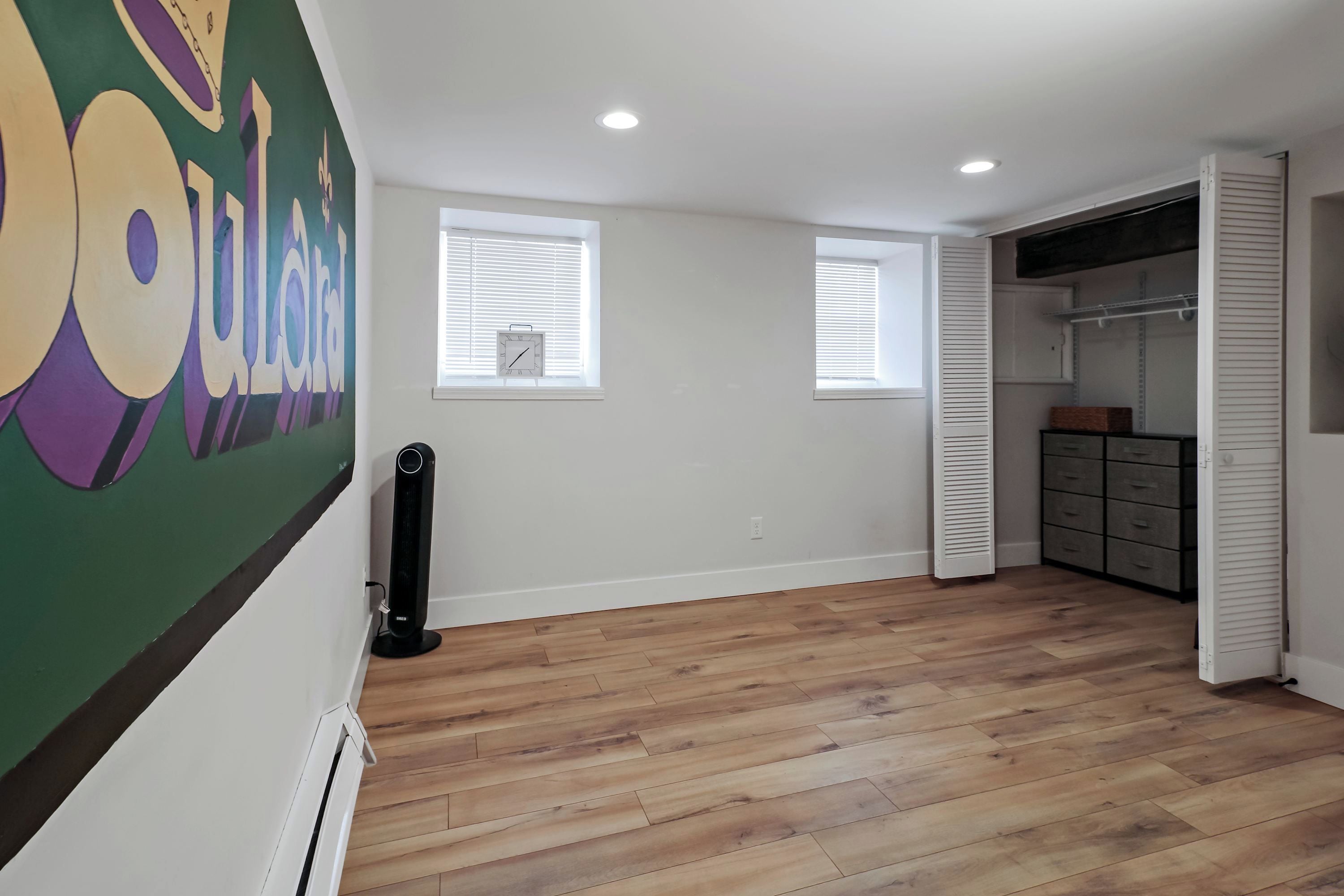 A room with wooden flooring, a large wall mural, two windows with blinds, a closet with shelves, and a radiator.