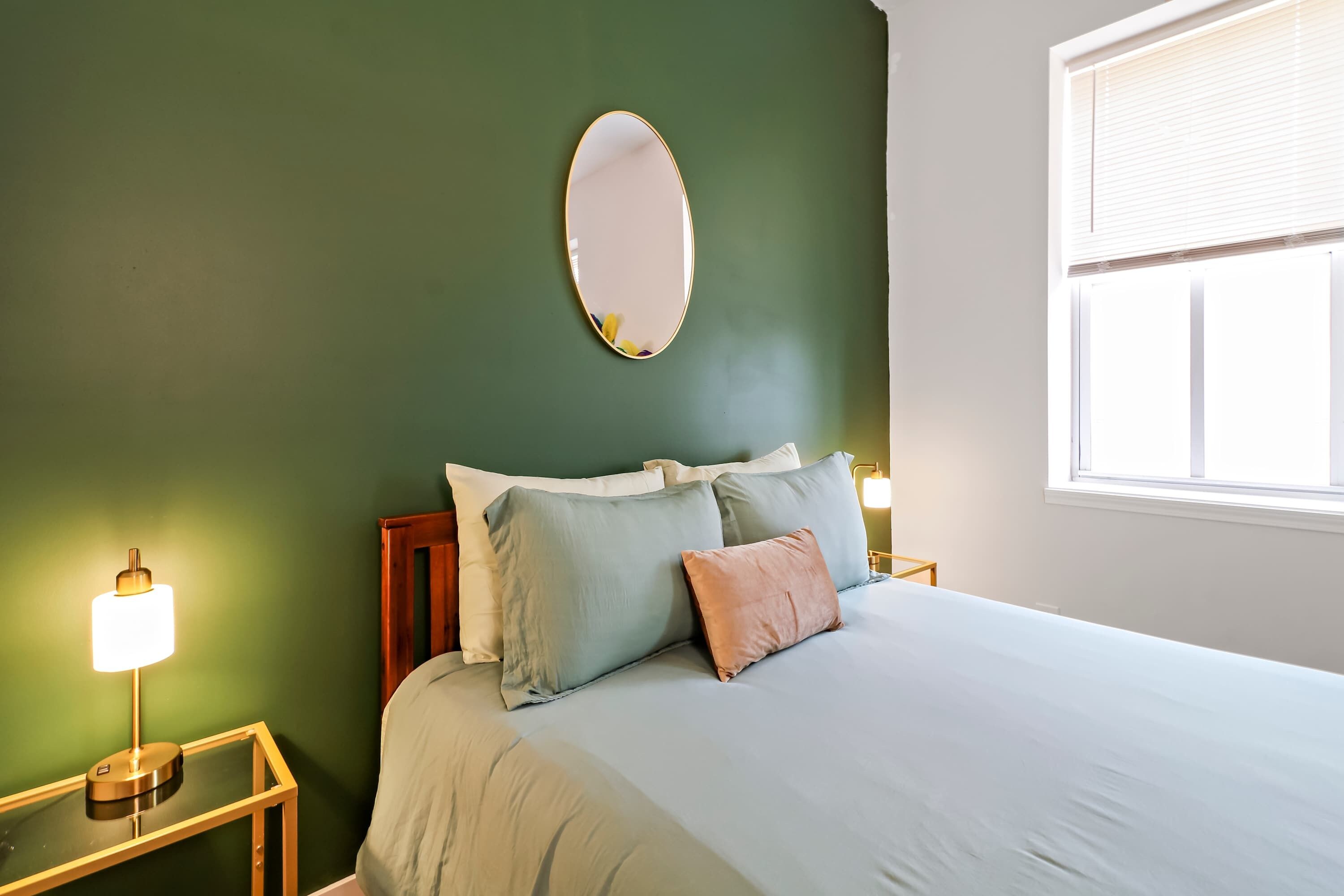 A bedroom with a bed, a bedside table with a lamp, a mirror on the wall, and a window.