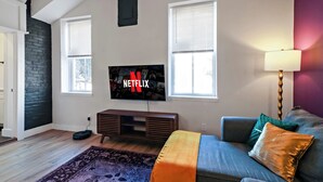Sala de estar com televisão de tela plana na parede, sofá com almofadas, luminária e tapete.