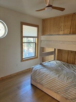 Une chambre avec un lit superposé, un ventilateur de plafond, une fenêtre avec des stores et une vue sur les arbres à l’extérieur.