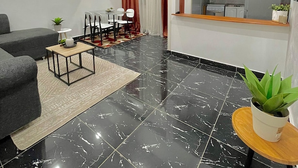 A modern living room with a black marble floor, a grey sofa, a wooden coffee table, and a dining area with chairs and a table. - Appartements VEKA (Pointe-Noire)