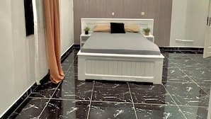 A modern bedroom with a bed, curtains, a nightstand, a potted plant, and a ceiling light fixture. - Appartements VEKA (Pointe-Noire)