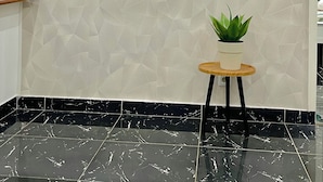 A room with a flat-screen TV mounted on a textured wall, a small wooden table with a potted plant, and black marble flooring. - Appartements VEKA (Pointe-Noire)