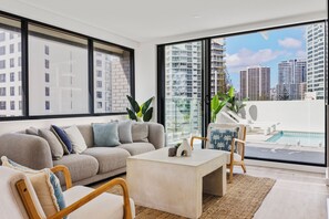 A modern living room with a large window, a sofa, a coffee table, and a view of a swimming pool and high-rise buildings. - The Shores Surfers Paradise Apartments (Surfers Paradise)