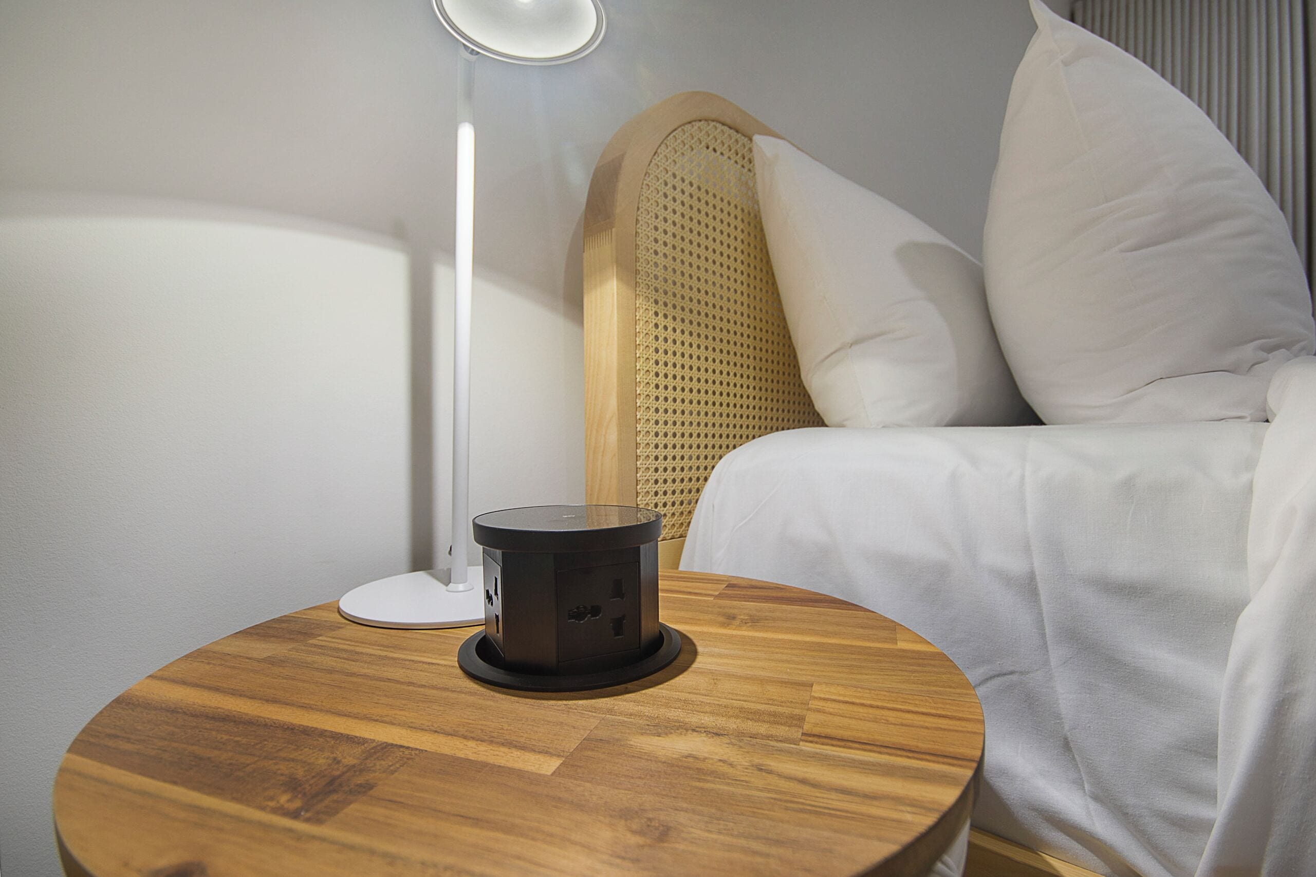 Een net opgemaakt bed met witte beddengoed, een rond houten nachtkastje met een zwart voorwerp erop, en een witte vloerlamp.