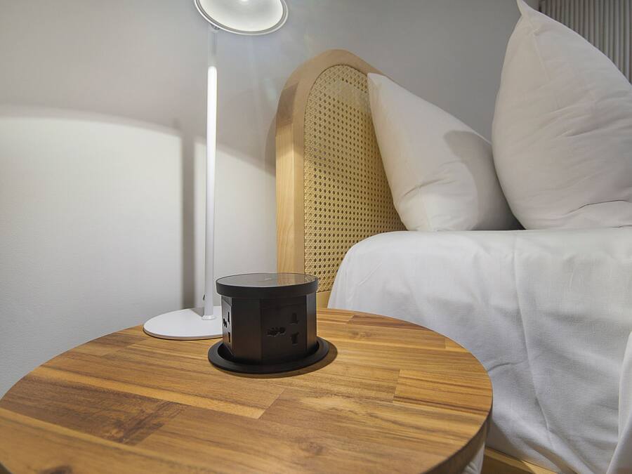 Een net opgemaakt bed met witte beddengoed, een rond houten nachtkastje met een zwart voorwerp erop, en een witte vloerlamp.