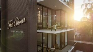 Een modern gebouw met een glazen balkon, een bord met de tekst 'The Shores - Surfers Paradise' en uitzicht op palmbomen.