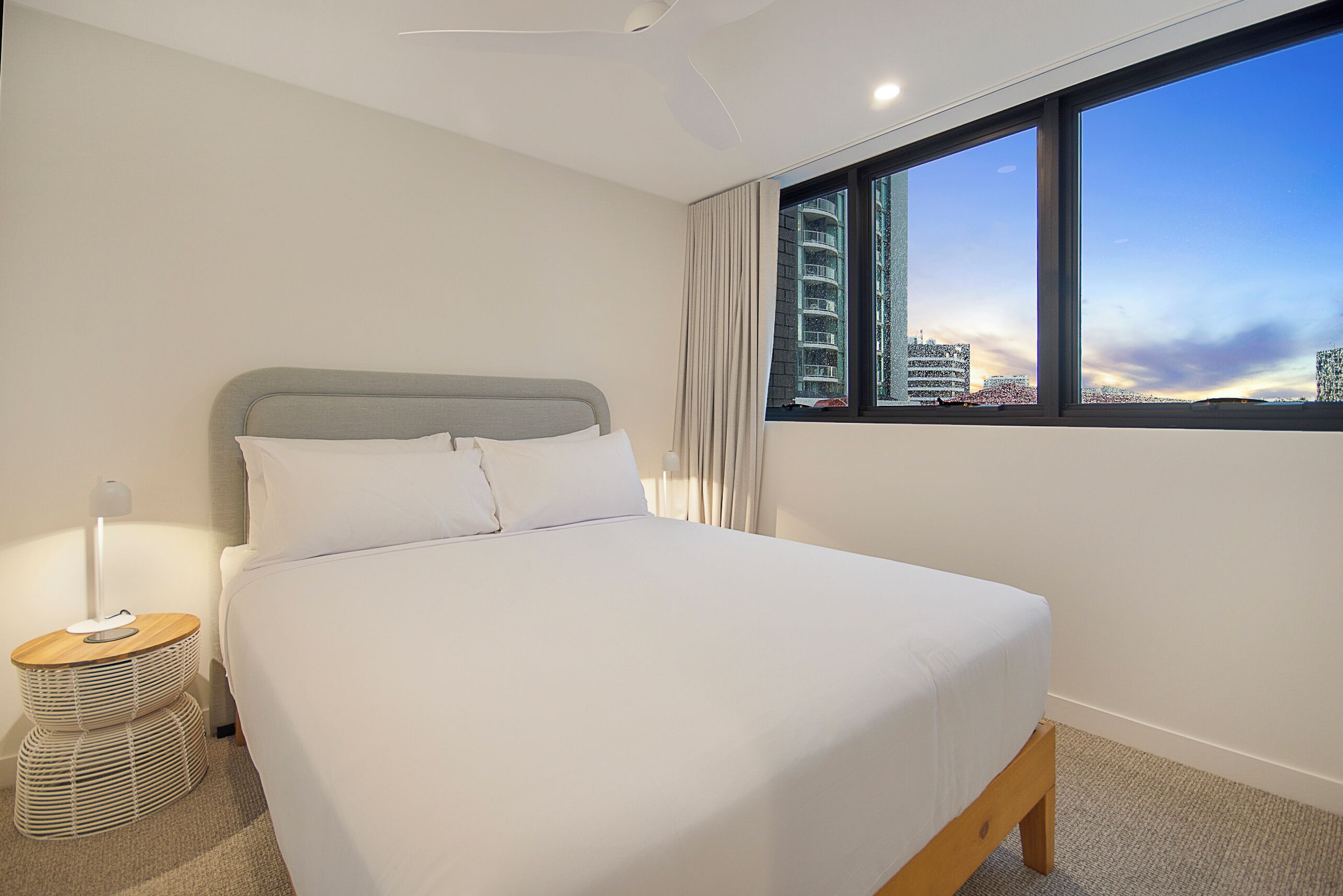 Una habitación de hotel moderna con una cama grande, una mesita de noche con lámpara y vistas a la ciudad a través de un amplio ventanal.