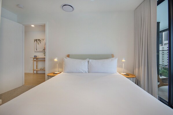 Habitación de hotel moderna con una cama grande, dos mesitas de noche con l ámparas, vista de la ciudad a través de un ventanal y una puerta que da a un pasillo con suelo de madera y un estante con objetos decorativos.