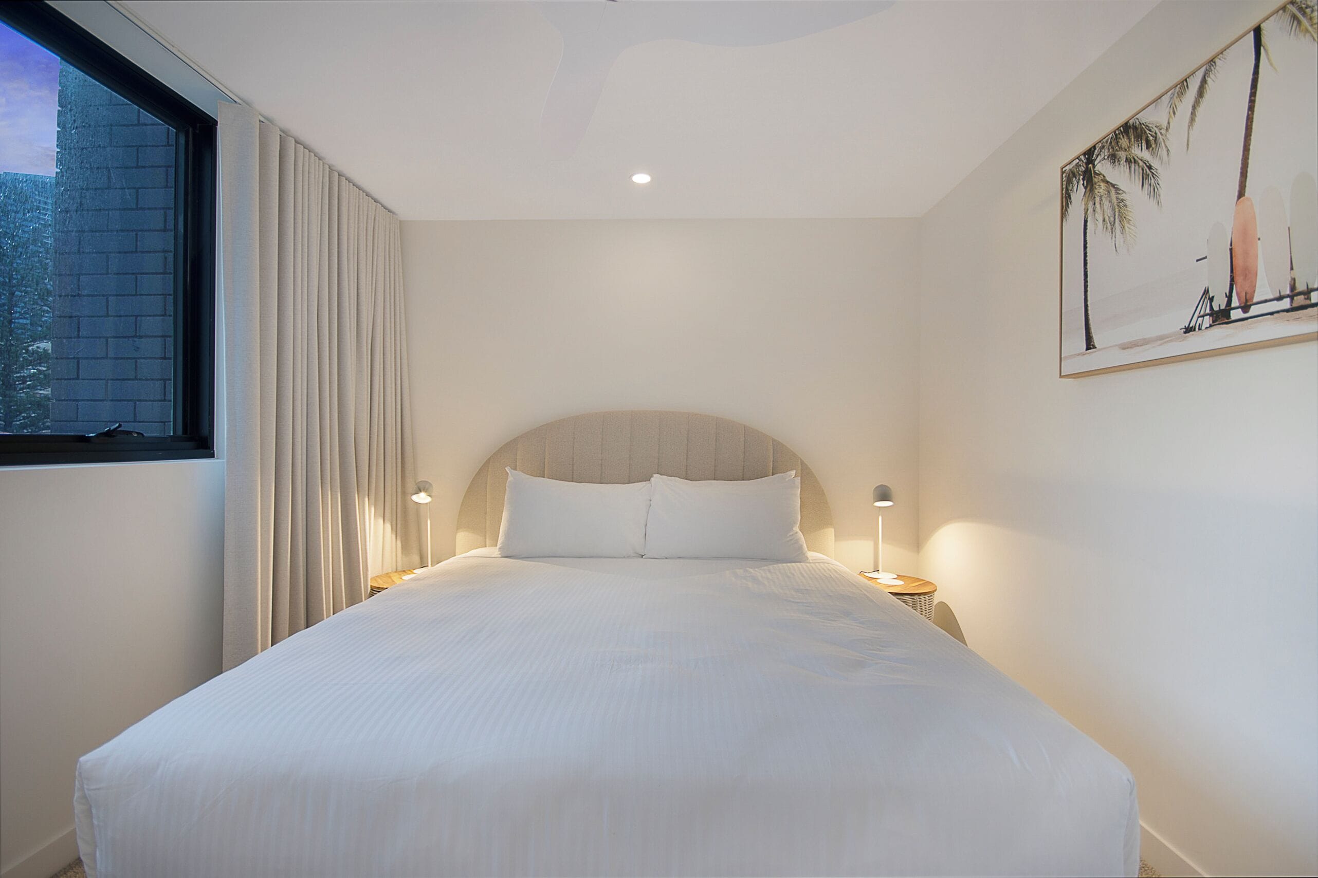 A hotel room with a large bed, a window with curtains, a headboard, and a wall-mounted artwork.