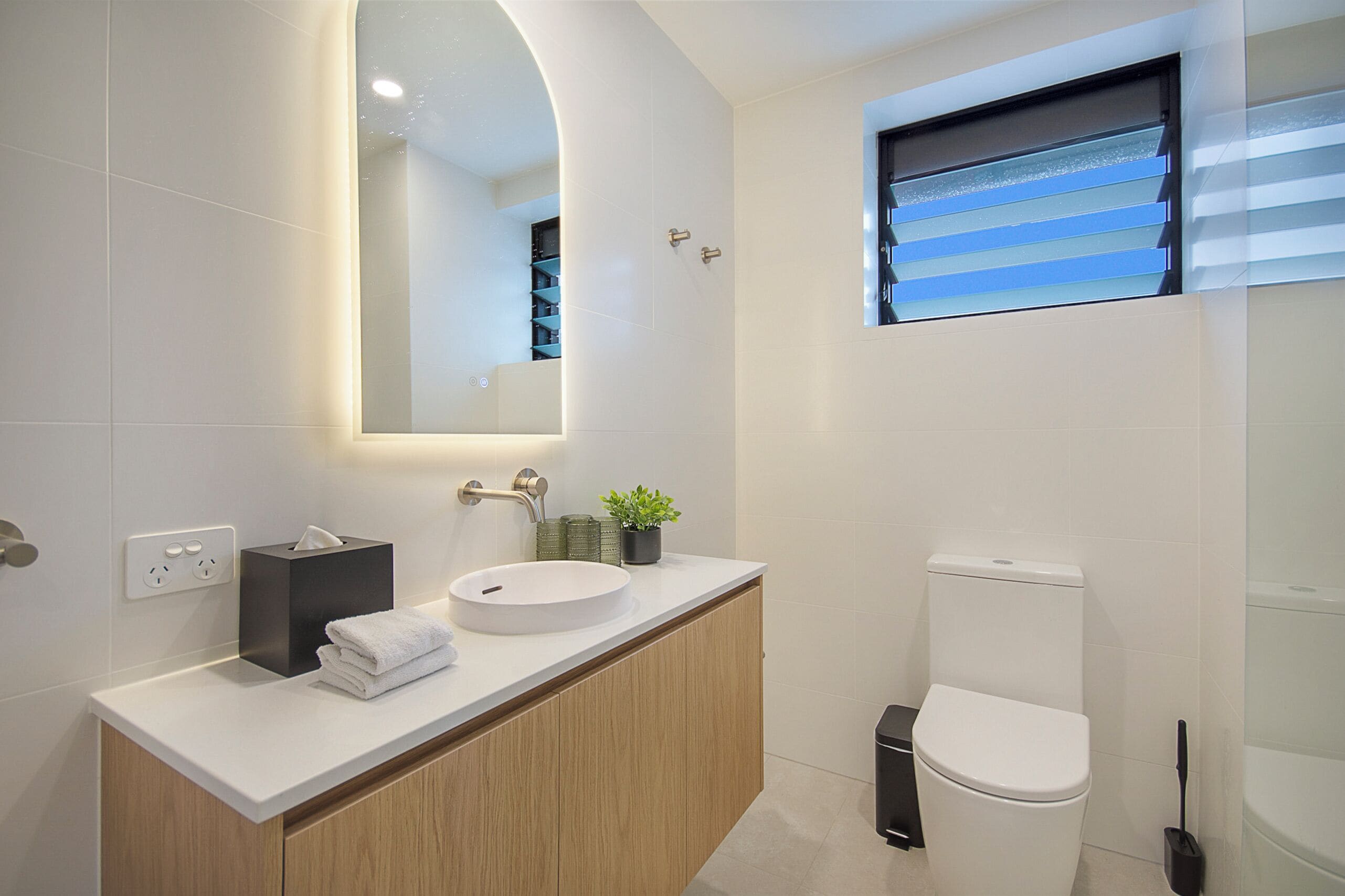 Une salle de bain moderne avec un lavabo blanc, un miroir, des toilettes et une fenêtre avec des stores.