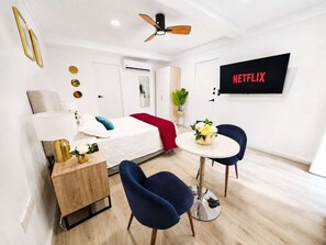Une chambre d’hôtel moderne équipée d’un lit, d’une table de chevet, de chaises, d’une petite table avec un vase, d’un ventilateur de plafond et d’un téléviseur fixé au mur qui affiche « NETFLIX ».