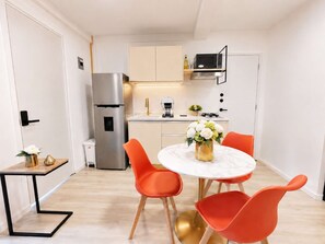 Une cuisine compacte équipée d’un réfrigérateur, d’une machine à café et d’un four à micro-ondes. On y trouve une table à manger avec deux chaises et un vase de fleurs.
