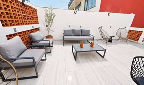 Un área de estar moderna al aire libre con un sofá, sillas y una mesa de centro, que presenta un diseño geométrico en la pared y plantas en macetas.