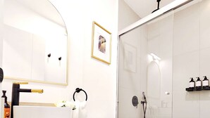 A modern bathroom with a white sink, toilet, and shower. There is a framed picture on the wall and a shelf with toiletries.