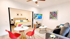 A modern hotel room with a bed, a TV, a sofa, and a dining area with red chairs and a table.