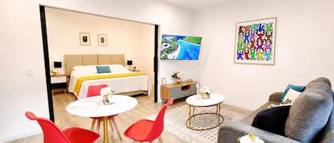 A modern hotel room with a bed, a TV, a sofa, and a dining area with red chairs and a table.