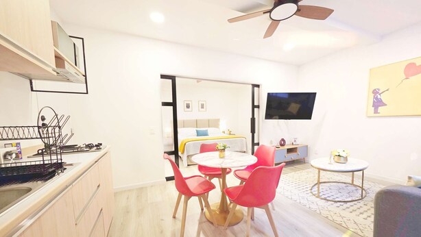 A modern kitchen with a built-in stove, a dining area with a round table and red chairs, a ceiling fan, and a TV mounted on the wall.