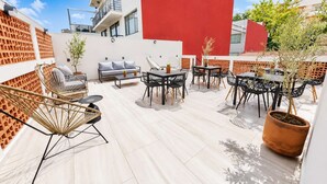 Un patio exterior con disposición de asientos, incluyendo sofás y mesas, rodeado por un muro blanco con una sección roja.