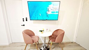A room with a TV mounted on the wall displaying a coastal view, two pink chairs around a small white table, and a wooden floor. - Chic Polanco Loft: Prime Spot for Urban Explorers (Ciudad de México)
