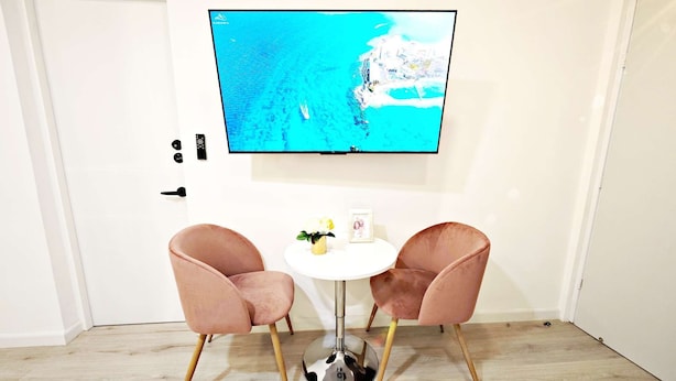 Una habitación con un televisor en la pared que muestra una vista costera, dos sillas rosas alrededor de una pequeña mesa blanca y un suelo de madera.