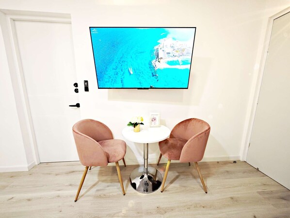A room with a TV mounted on the wall displaying a coastal view, two pink chairs around a small white table, and a wooden floor. - Chic Polanco Loft: Prime Spot for Urban Explorers (Ciudad de México)