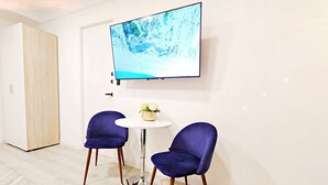 Ein modernes Zimmer mit einem an der Wand befestigten Flachbildfernseher, zwei blauen Stühlen um einen kleinen weißen Tisch mit Blumenvase und einer Holztür.