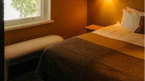 Une chambre avec un lit simple, un banc, une fenêtre donnant sur des arbres et un luminaire fixé au mur.