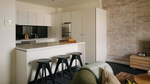 A modern kitchen with white cabinetry, a stainless steel refrigerator, and a black stove. There are three black stools at the counter. A brick wall is visible in the background. - Gibson's Mill 3 Bedroom Loft Penthouse with Private
Balcony&Waterfront Views-301 (Hobart)
