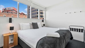 Habitación de hotel moderna con una cama grande, una mesita de noche con lámpara y vistas a edificios a través de la ventana.