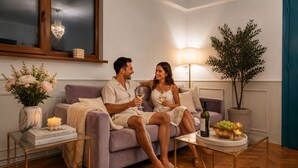 Una pareja sentada en un sofá con una lámpara, un jarrón de flores y un libro titulado 'LIVE BEAUTIFUL' sobre una mesita.