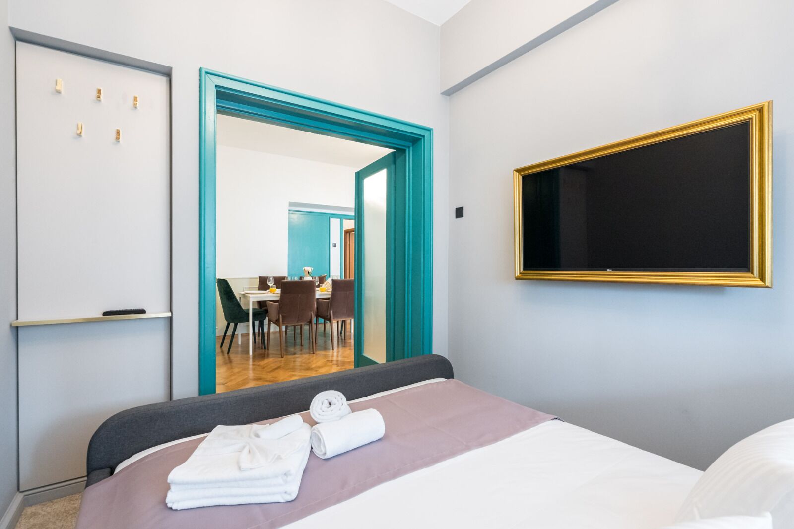 Habitación de hotel con cama, televisor, escritorio con sillas y un área de comedor visible a través de una puerta abierta.