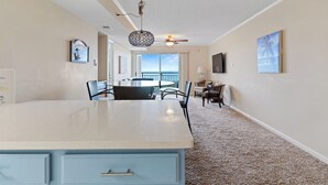 A spacious living area with a large island table, a ceiling fan, and a view of the ocean through a sliding door.