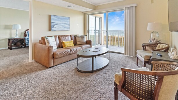 A spacious living room with a brown leather sofa, a round coffee table, and a wicker chair. A large window offers a view of the ocean.