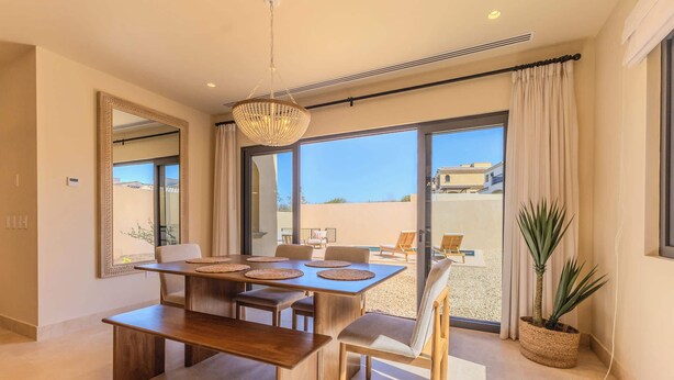 Un comedor con mesa y sillas de madera, una lámpara colgante y vistas al exterior a través de amplios ventanales.