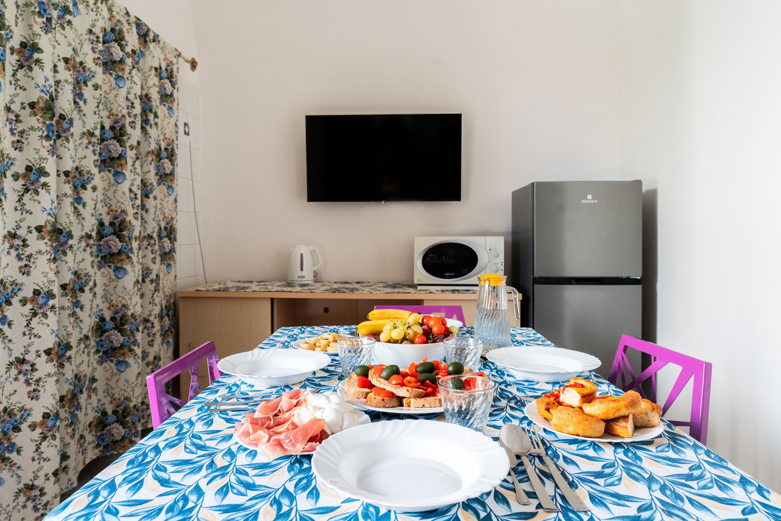 A dining table set with plates, cutlery, and food, a television mounted on the wall, and a refrigerator in the background.