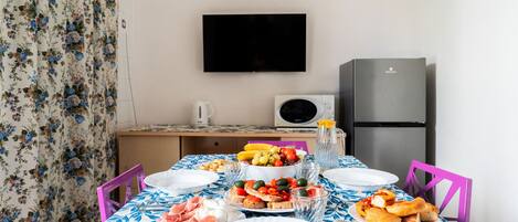 A dining table set with plates, cutlery, and food, a television mounted on the wall, and a refrigerator in the background.