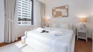 Une chambre d’hôtel comprenant un lit, deux tables de chevet, une fenêtre avec des rideaux, un tableau abstrait accroché au mur, et une vue sur un bâtiment à l’extérieur.