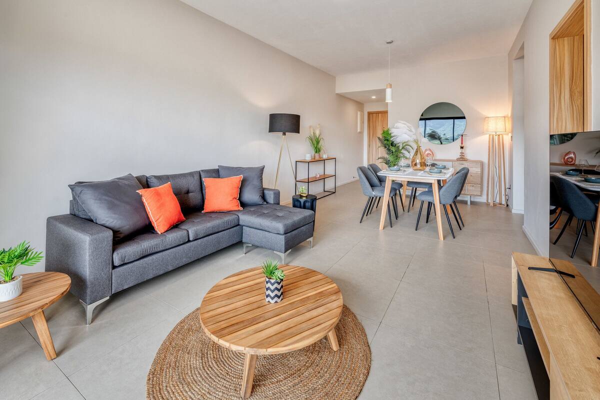 A modern living room with a grey sectional sofa, a round wooden coffee table, and a dining area with a white table and chairs.