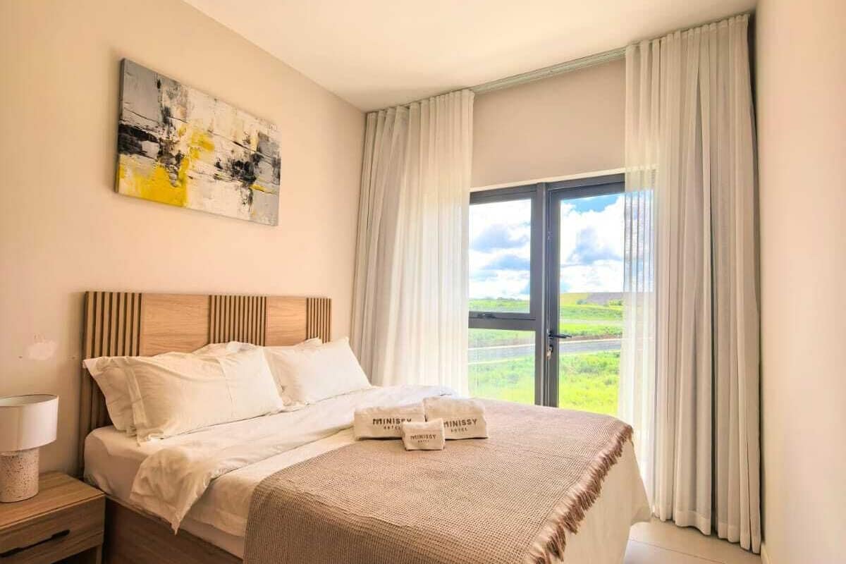 A bedroom with a bed, a nightstand, a lamp, a painting, and a view of the outdoors through a window.