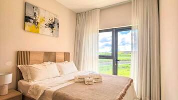 Un dormitorio con cama, mesita de noche, lámpara, un cuadro y vista al exterior a través de una ventana.