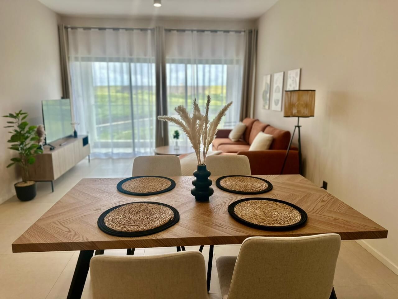 Uma mesa de jantar com quatro jogos americanos, um vaso com plantas secas e um sofá ao fundo.