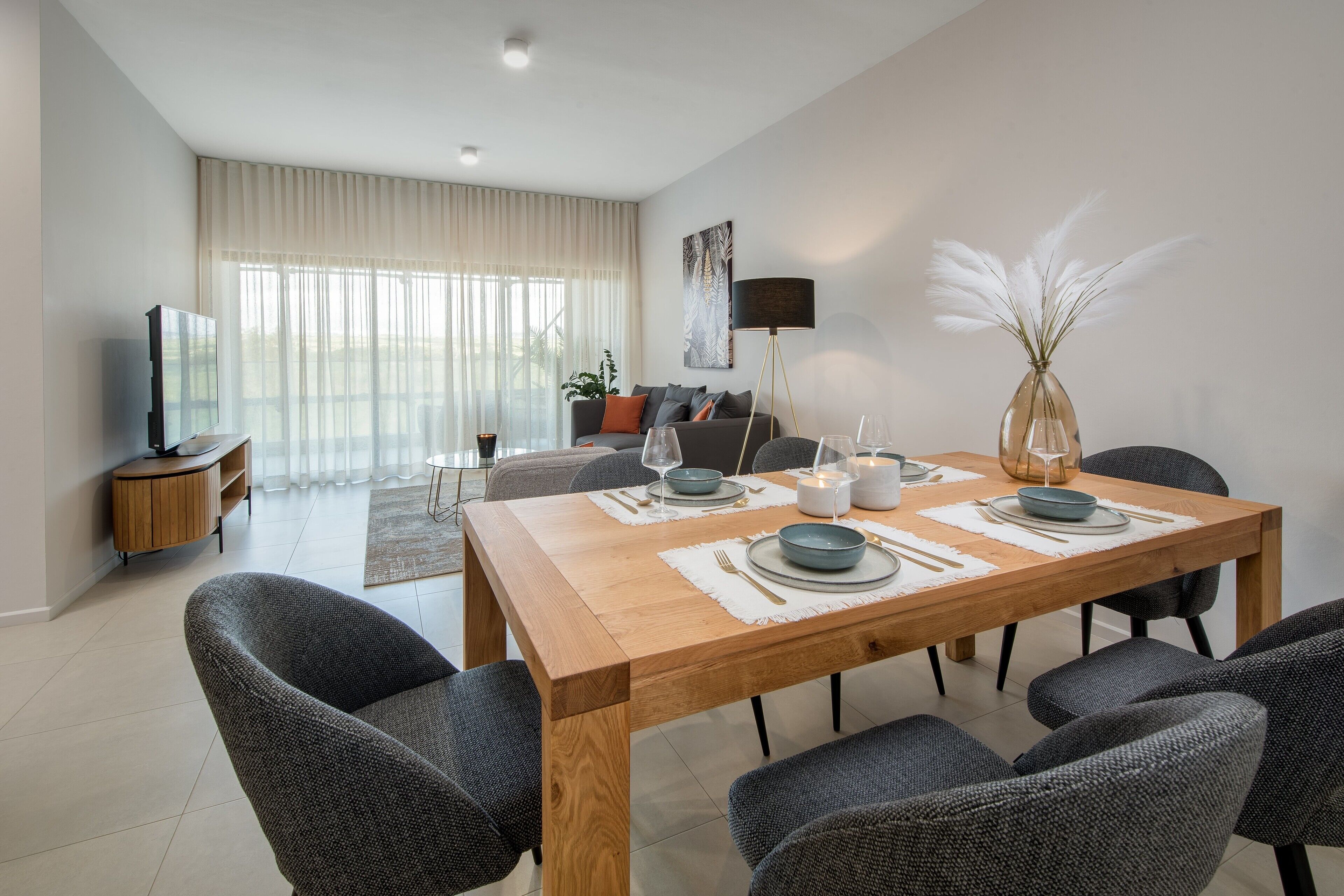 Una sala moderna con una mesa de comedor de madera dispuesta para cuatro personas, un sofá, un televisor y una lámpara de pie.